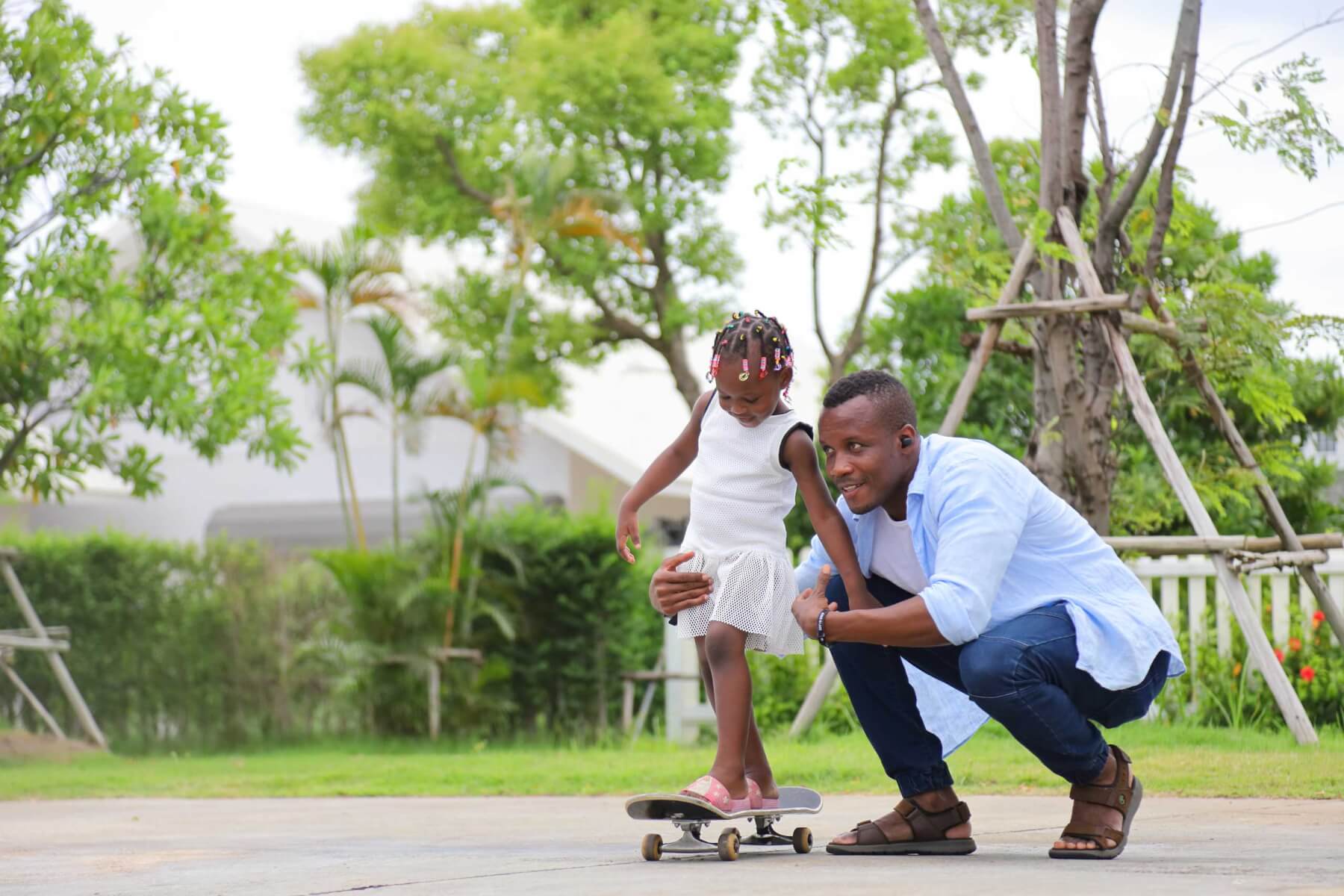 African American father teach his little daughter to play skateboard in the public park