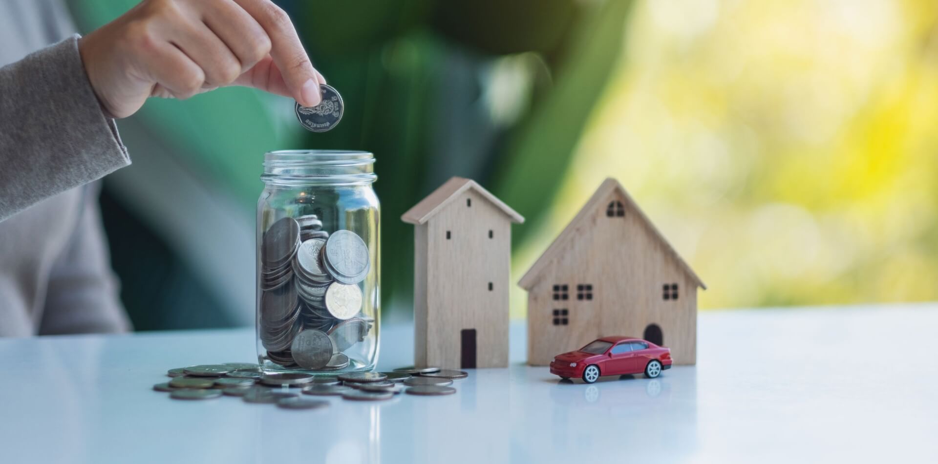 Closeup image of a woman collecting and putting coins in a glass jar with wooden house models and car figure model for saving money concept